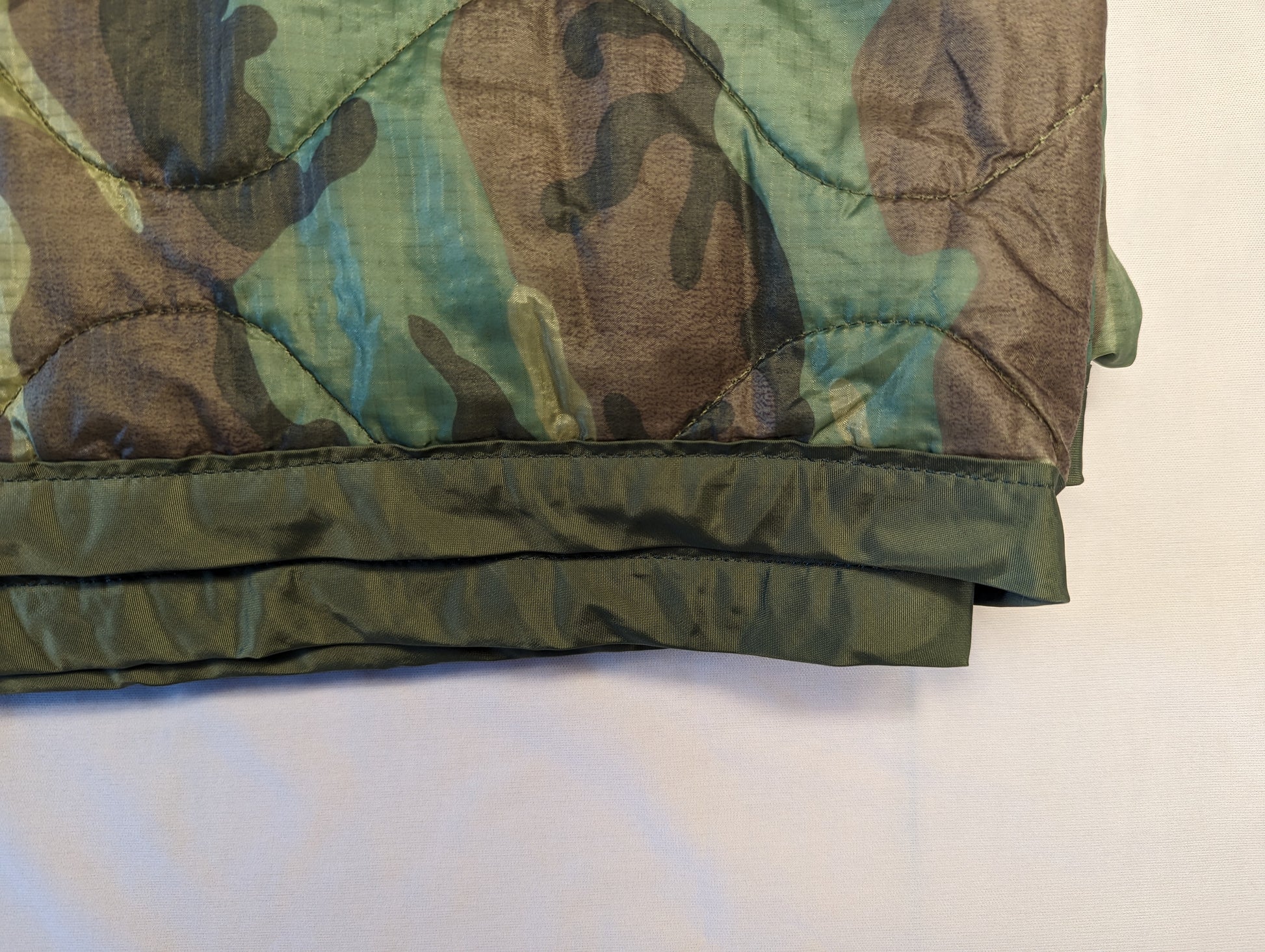 A close-up view of the bottom edge of a U.S. Military Woodland camouflage Poncho Liner (Woobie), highlighting the quilted fabric texture and the heavy, dark green binding.