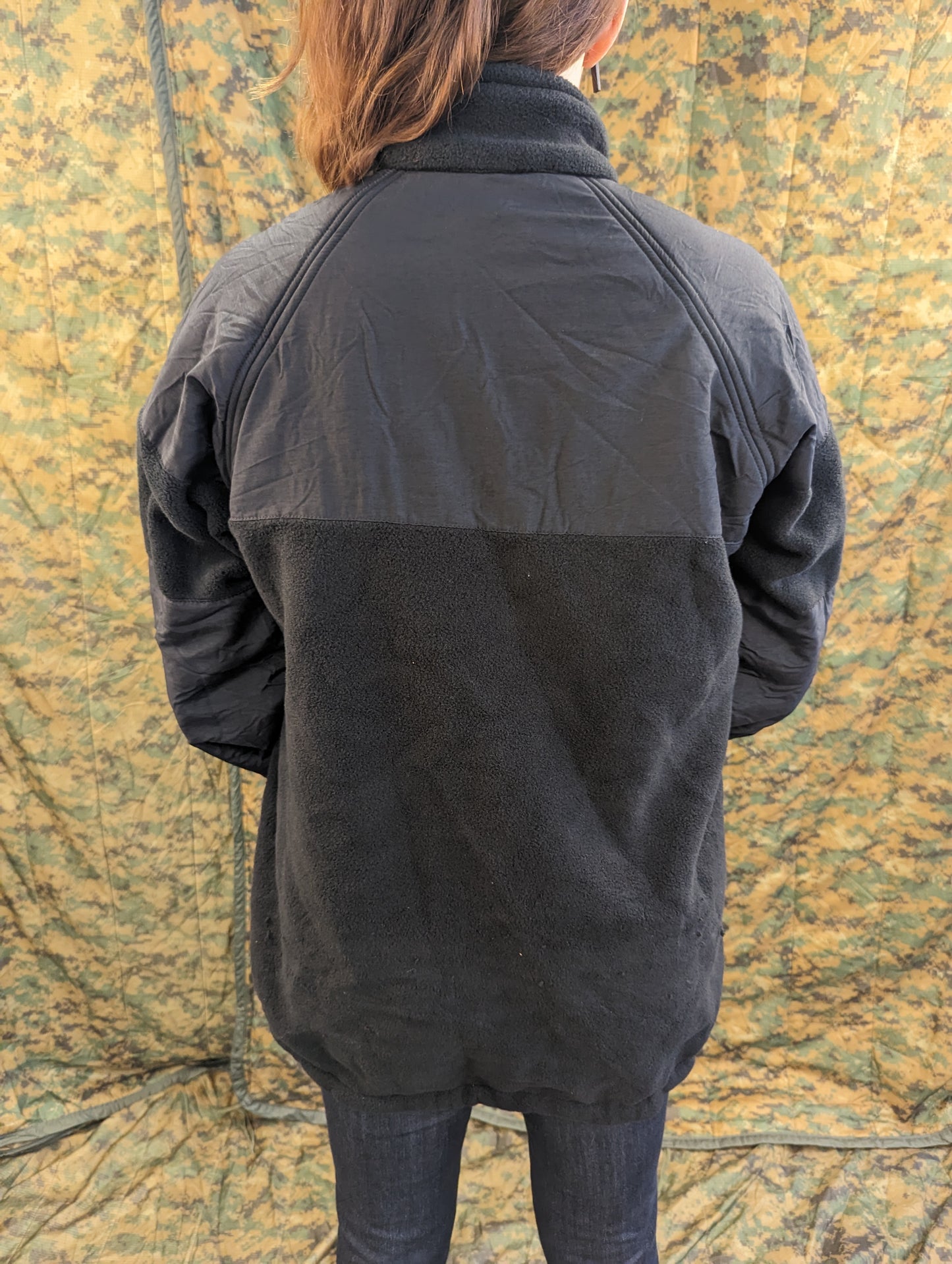 A back view of a person wearing a black U.S. Navy parka fleece liner, highlighting the smooth nylon yoke and the textured fleece material against a camouflage background.