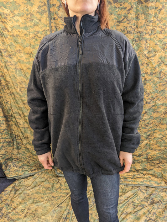 A person standing against a camouflage background, wearing a black U.S. Navy parka fleece liner, zipped up and showing the textured material and high collar design.