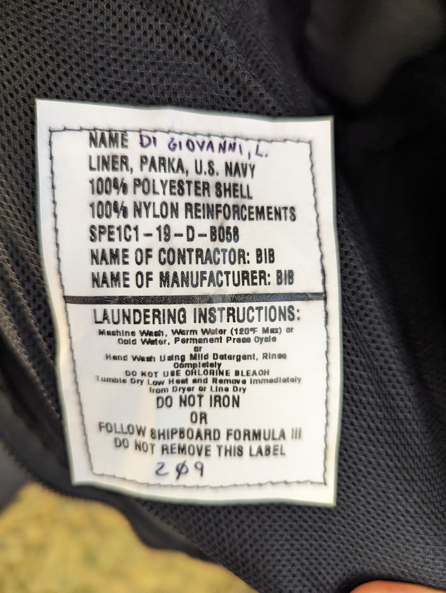 A close-up photograph of the U.S. Navy Parka Liner issue tag, detailing the material composition, laundering instructions, and featuring a handwritten military identification number.