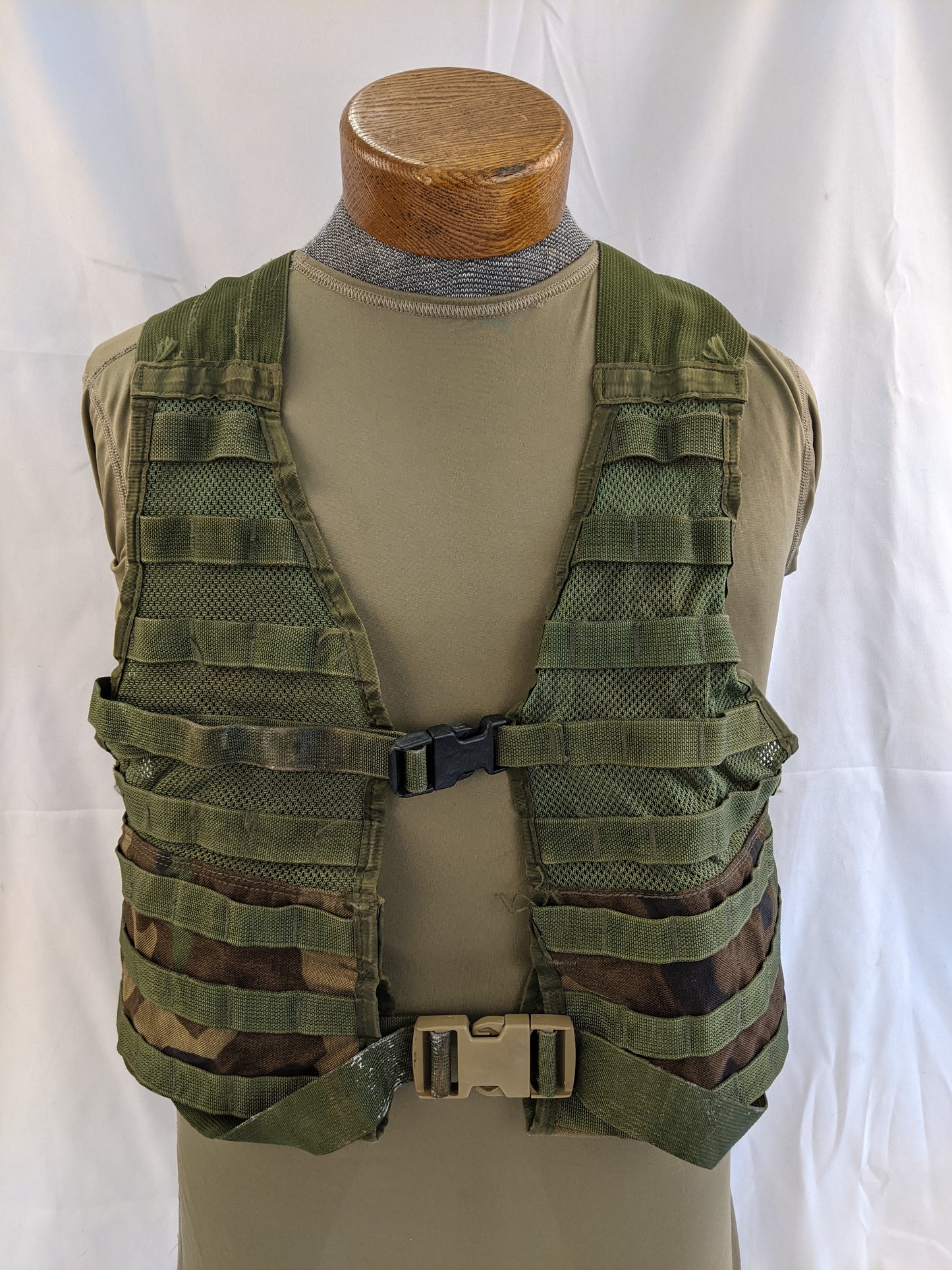 A front view of a U.S. Military Fighting Load Carrier (FLC) vest in Woodland camouflage pattern, displayed on a mannequin, highlighting the MOLLE webbing and quick-release buckles.