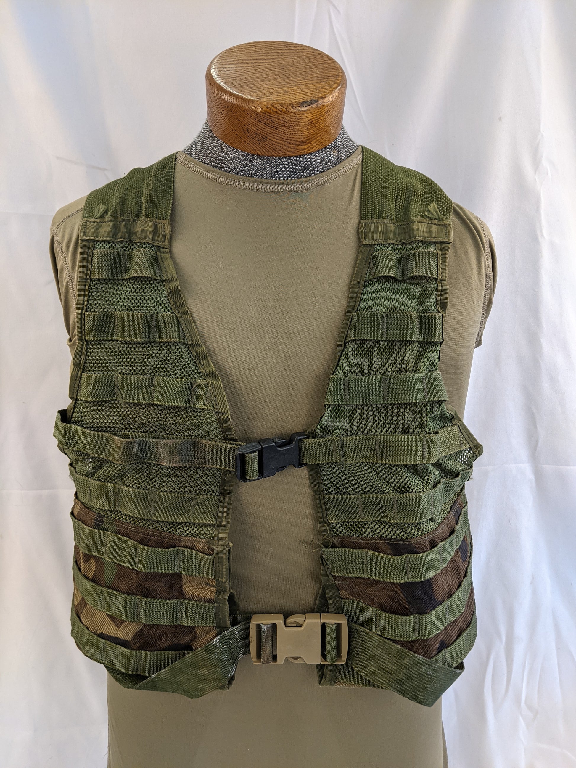 A front view of a U.S. Military Fighting Load Carrier (FLC) vest in Woodland camouflage pattern, displayed on a mannequin, highlighting the MOLLE webbing and quick-release buckles.