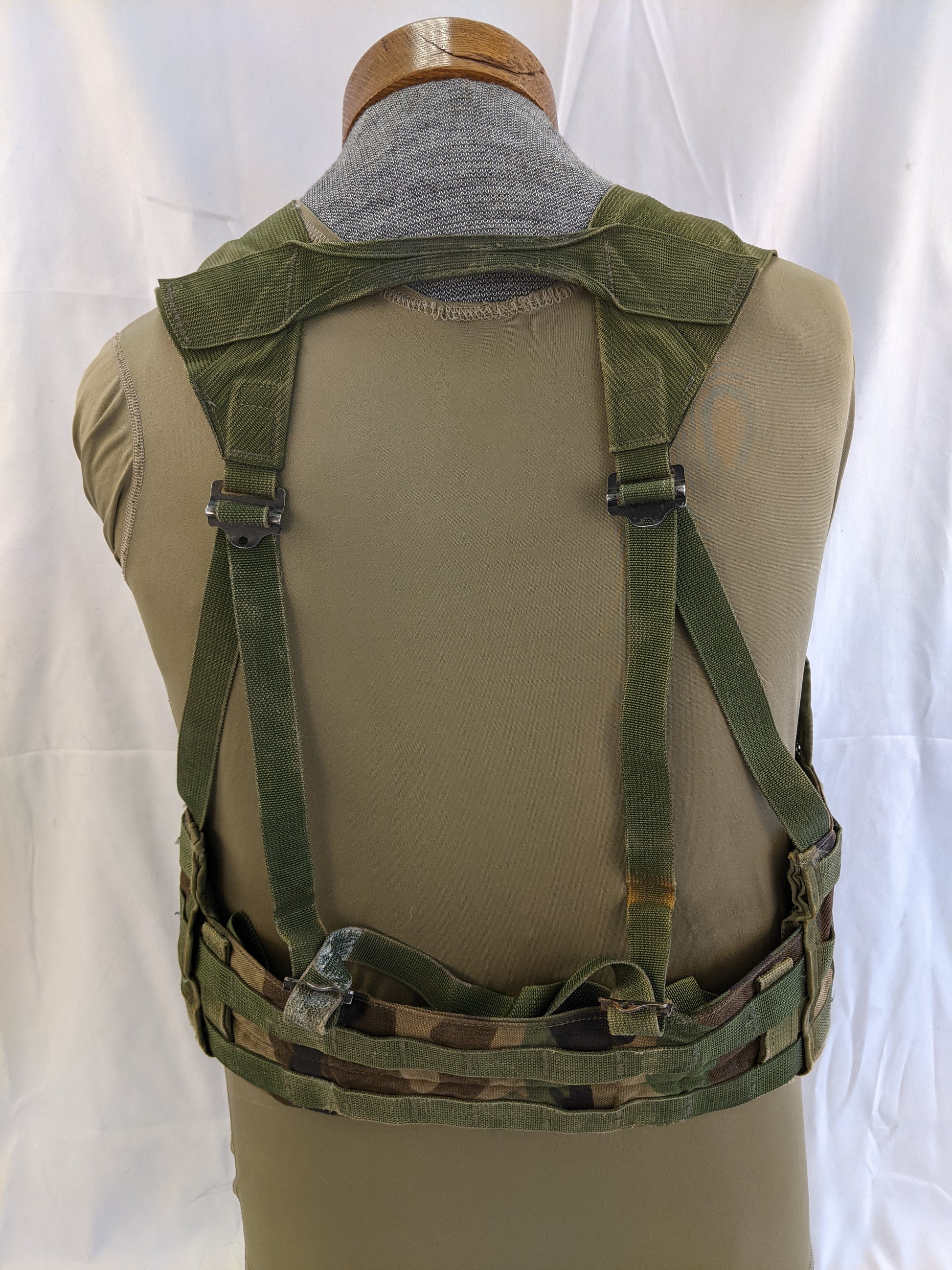A back view of a U.S. Military Fighting Load Carrier (FLC) vest in Woodland camouflage, mounted on a mannequin, highlighting the padded waist belt and adjustable shoulder harness.