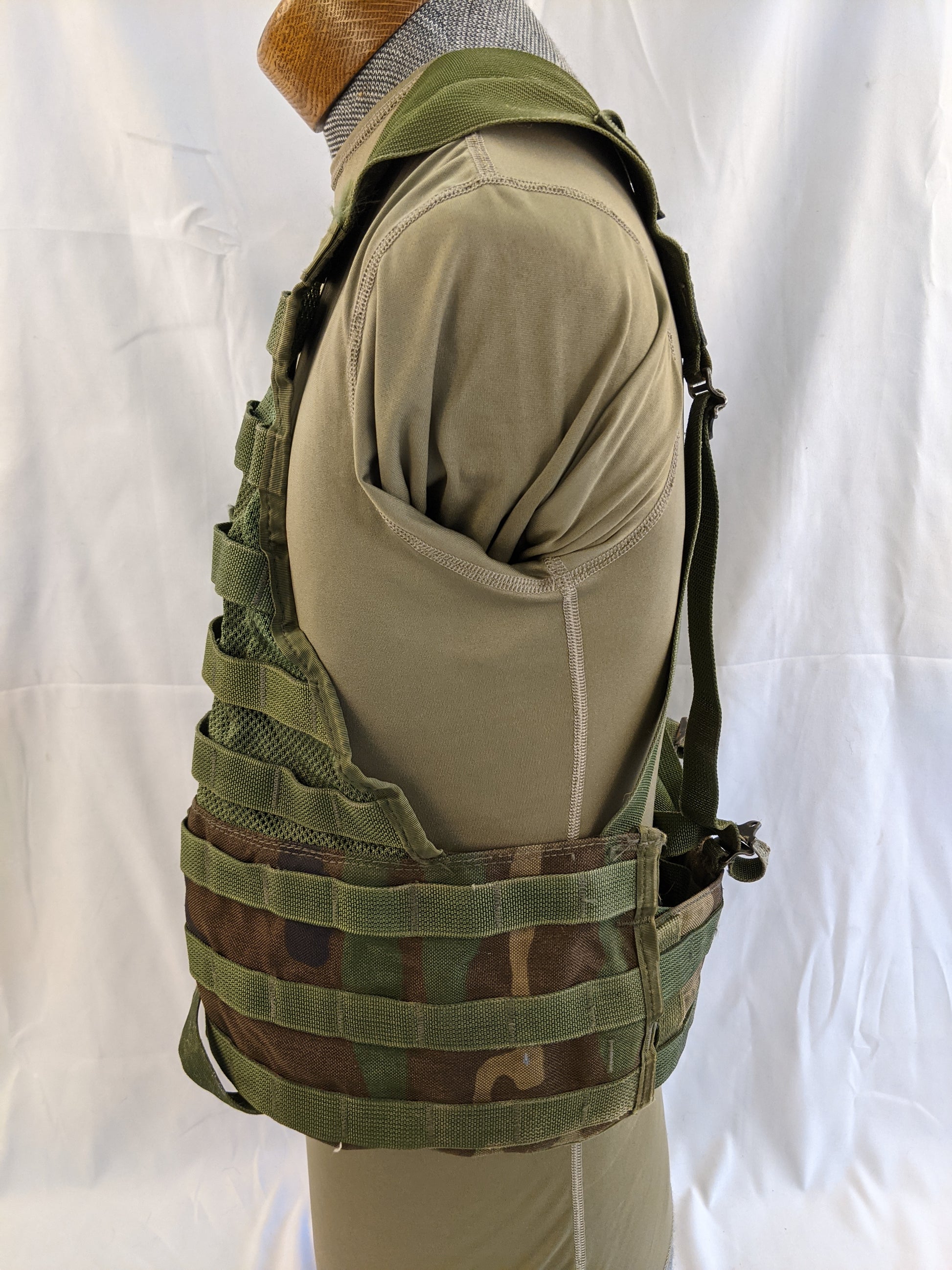 A side view of a U.S. Military Fighting Load Carrier (FLC) vest in Woodland camouflage pattern, mounted on a mannequin, highlighting the side MOLLE webbing and the shoulder strap attachment.