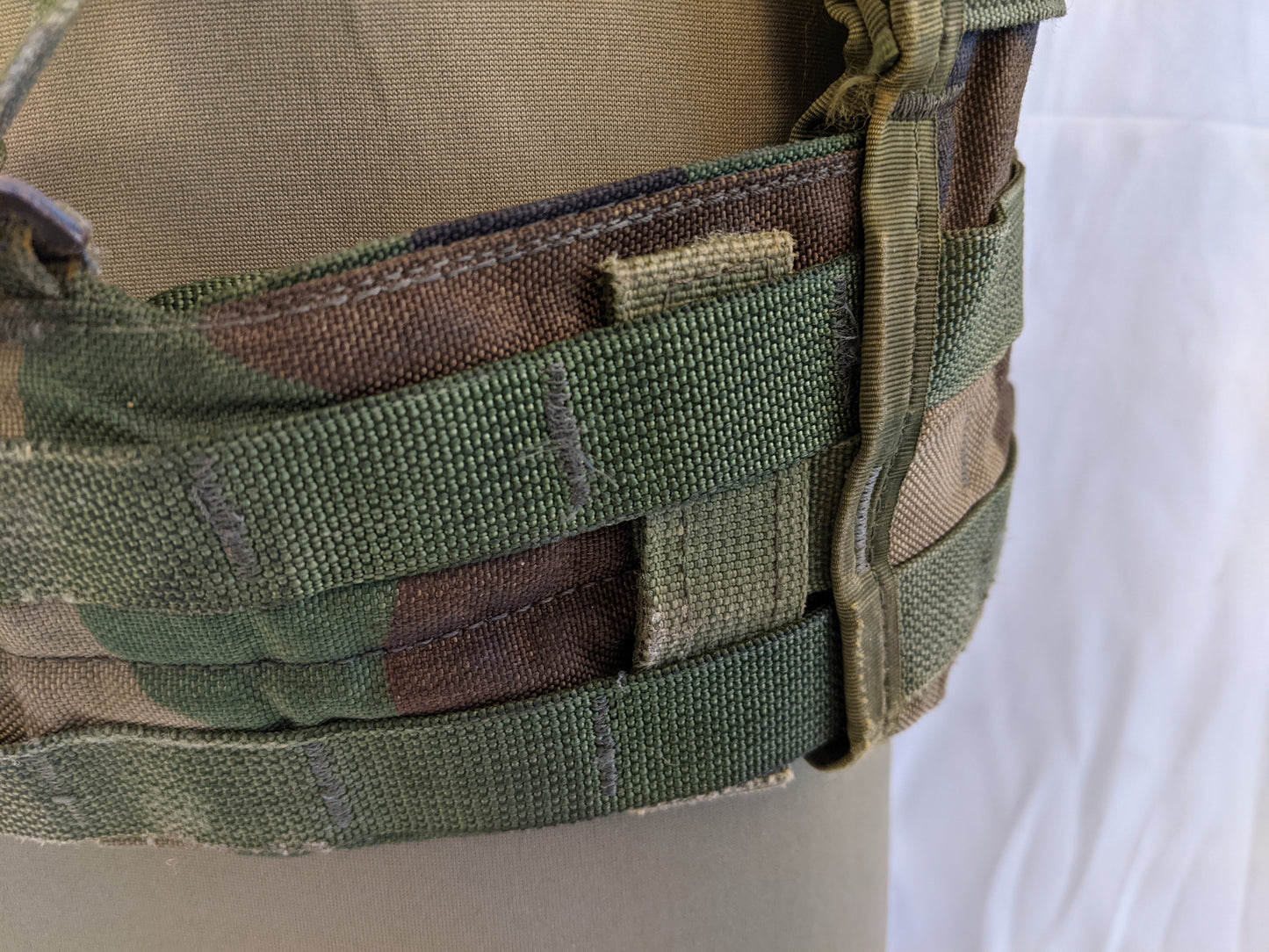 A close-up of the side panel of a U.S. Military Fighting Load Carrier (FLC) vest in Woodland camouflage, detailing the three rows of MOLLE webbing and the vertical adjustment strap.