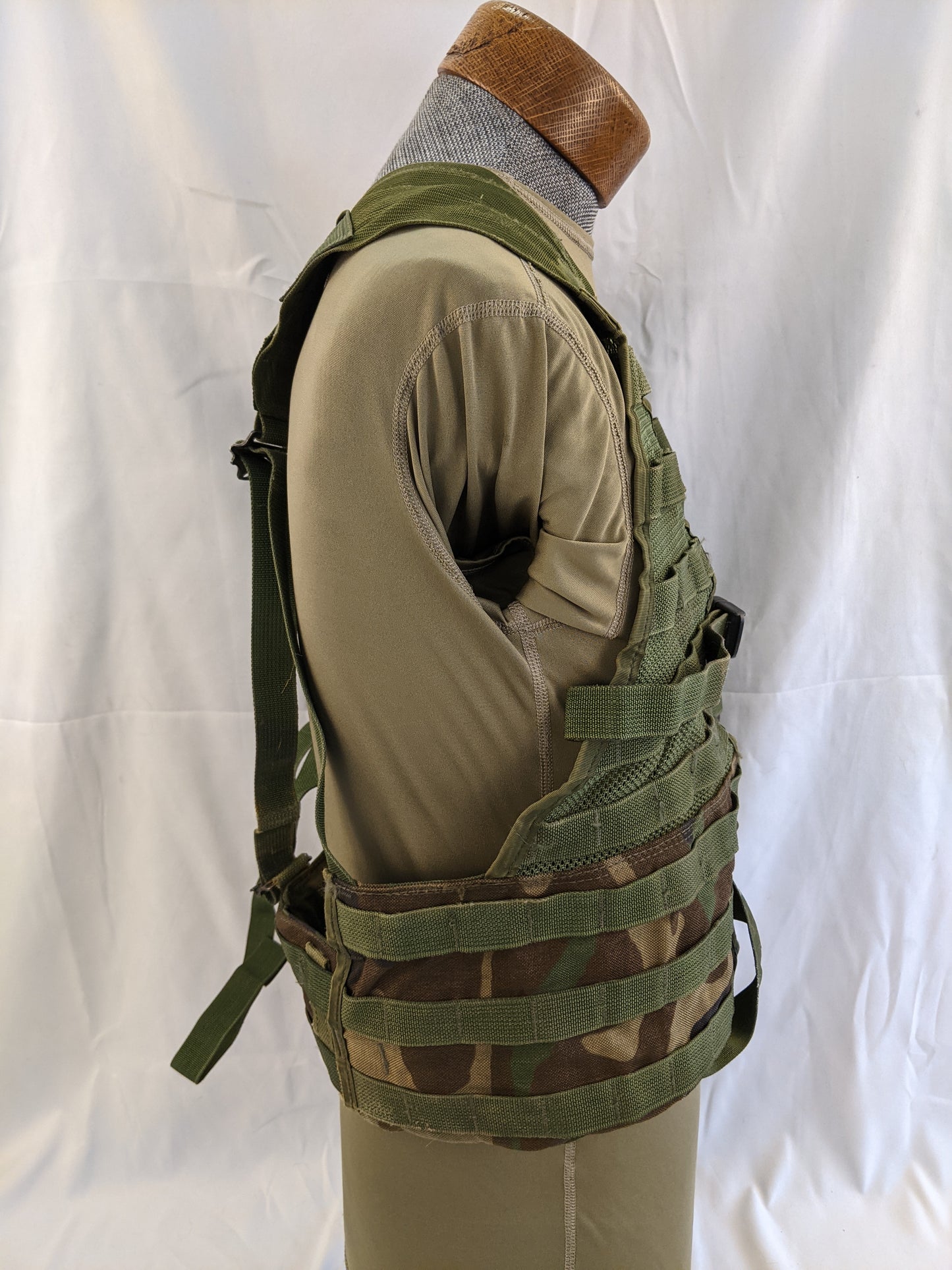 A side profile of a U.S. Military Fighting Load Carrier (FLC) vest in Woodland camouflage, mounted on a mannequin, detailing the MOLLE webbing and mesh backing on the torso panel.