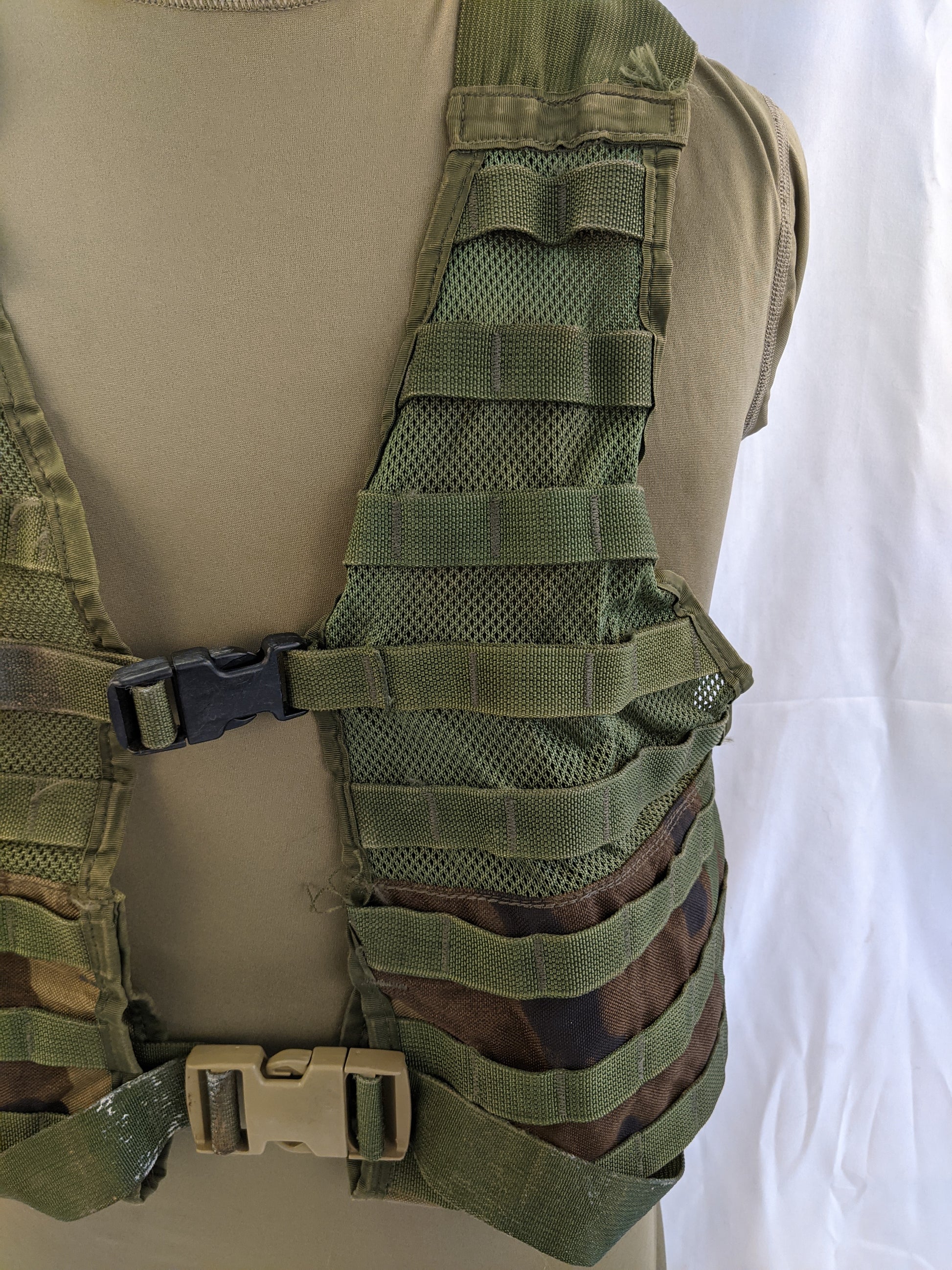 A close-up view of the U.S. Military Fighting Load Carrier (FLC) vest in Woodland camouflage, detailing the MOLLE webbing, the shoulder strap adjustment, and the quick-release chest buckle.