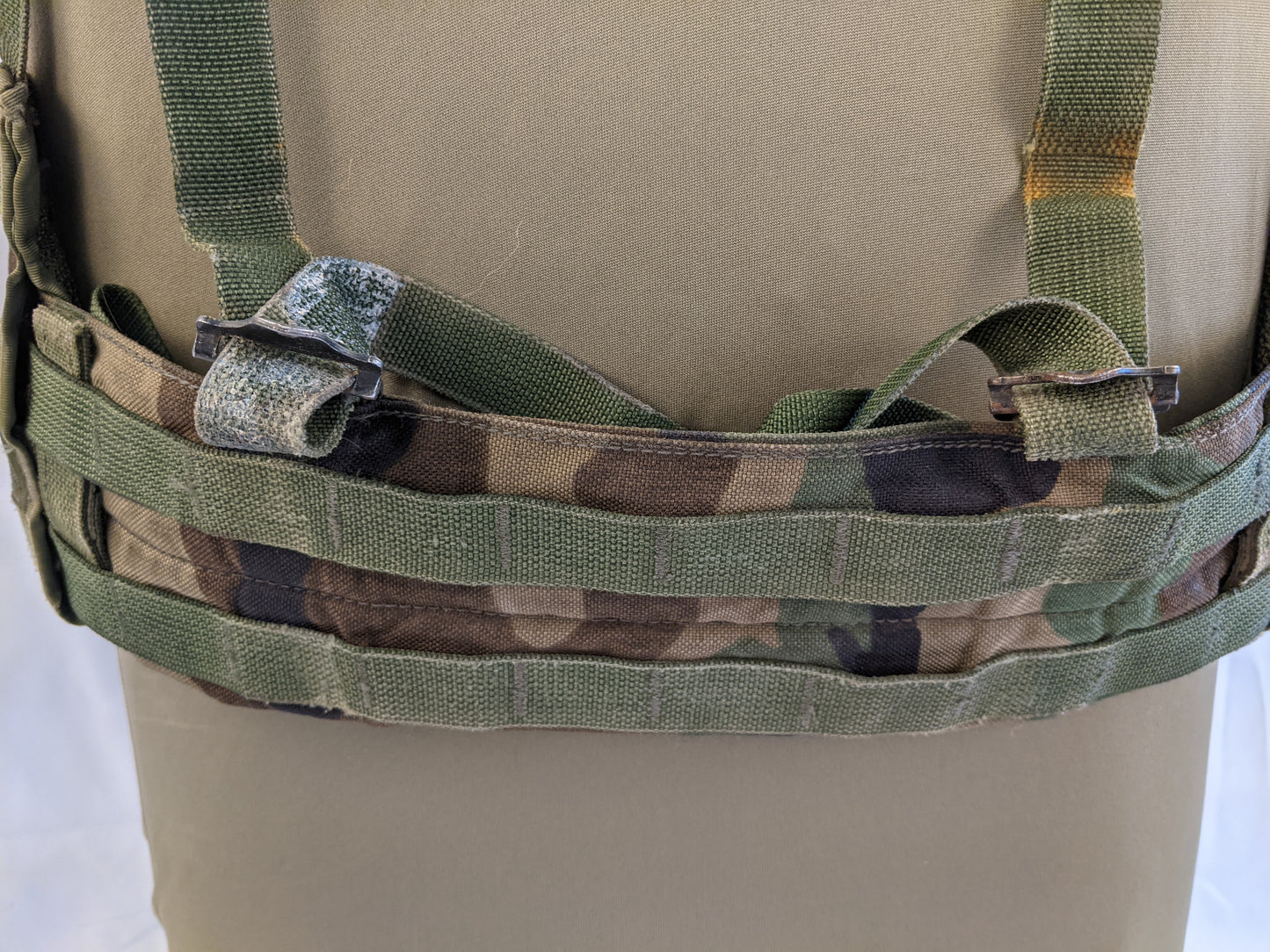 A close-up view of the U.S. Military Fighting Load Carrier (FLC) vest in Woodland camouflage, detailing the padded waist belt, the three rows of MOLLE webbing, and the connecting quick-release buckles.