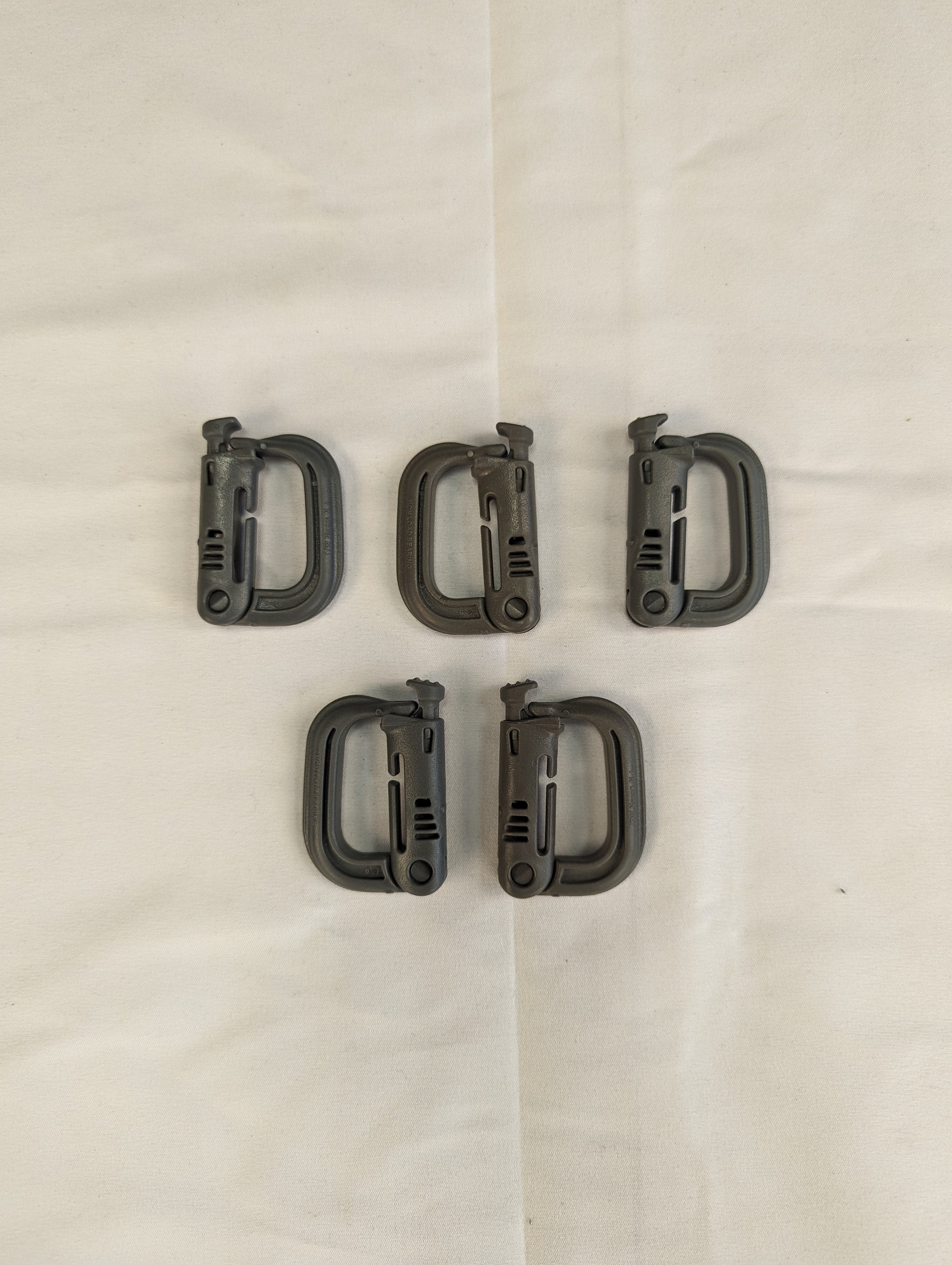 An overhead studio shot of five green ITW Nexus MOLLE Grimloc D-ring carabiner clips arranged in two rows on a wrinkled white fabric background.