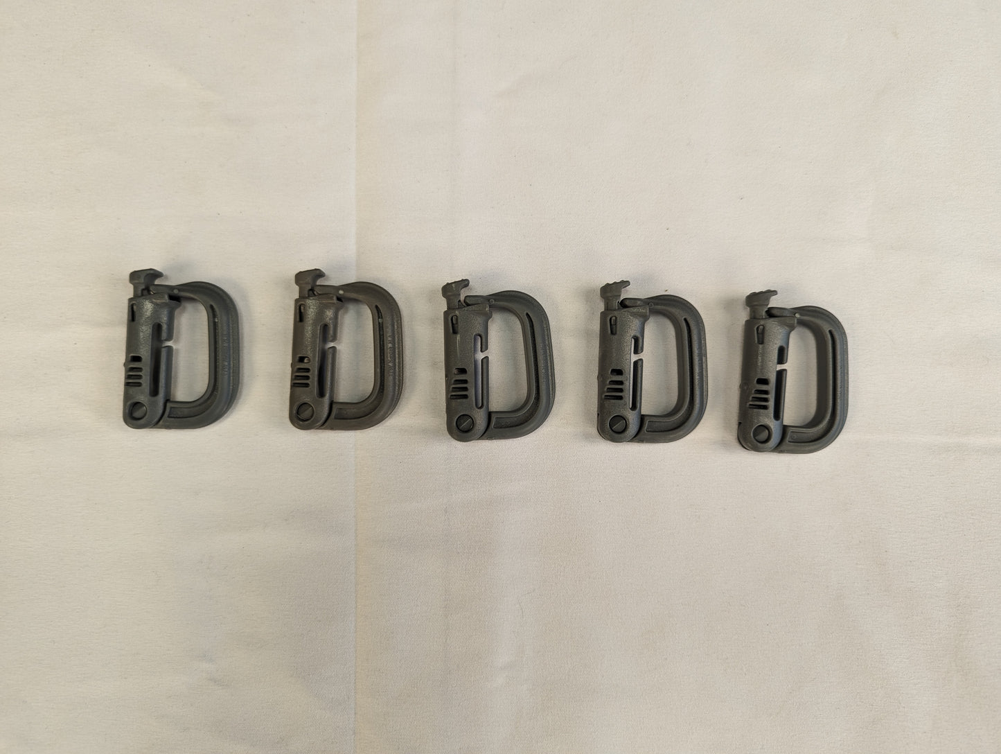 A close-up studio shot of five green ITW Nexus MOLLE Grimloc D-ring carabiner clips linked together in a chain, resting horizontally on a wrinkled white fabric background.