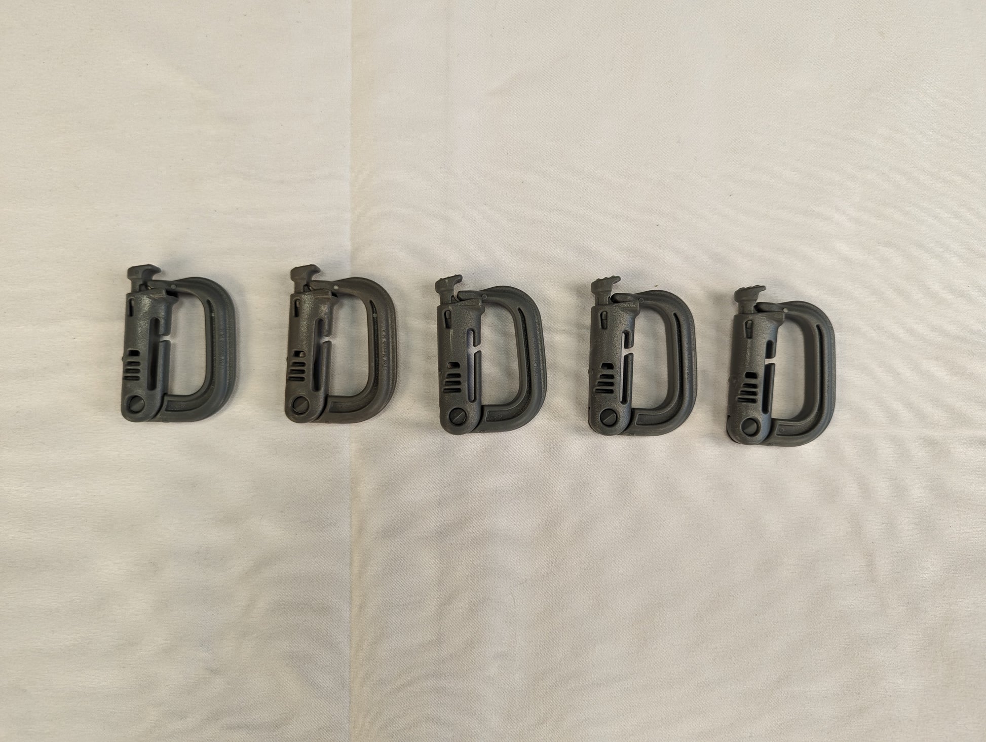 A close-up studio shot of five green ITW Nexus MOLLE Grimloc D-ring carabiner clips linked together in a chain, resting horizontally on a wrinkled white fabric background.