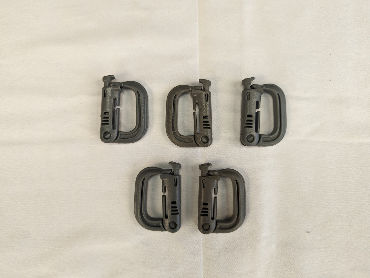An overhead studio shot of five green ITW Nexus MOLLE Grimloc D-ring carabiner clips arranged in two rows on a wrinkled white fabric background.