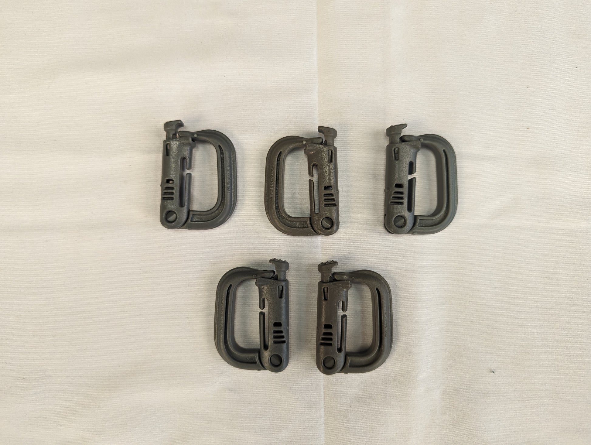 An overhead studio shot of five green ITW Nexus MOLLE Grimloc D-ring carabiner clips arranged in two rows on a wrinkled white fabric background.