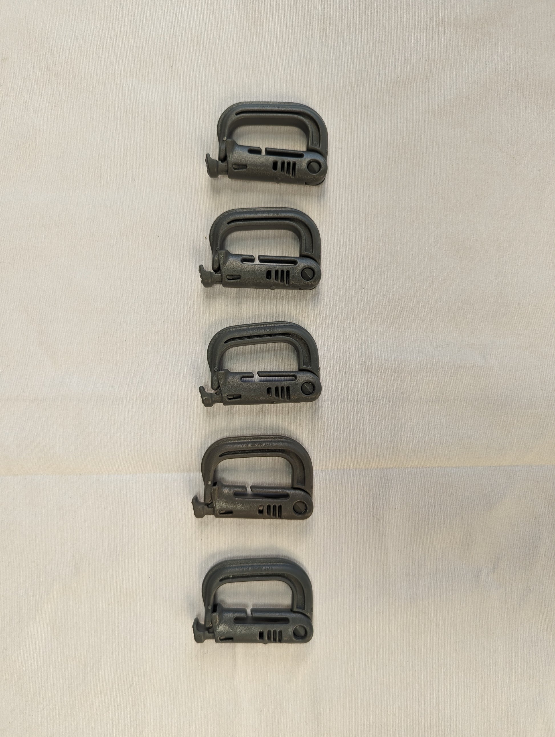 An overhead studio shot of five green ITW Nexus MOLLE Grimloc D-ring carabiner clips arranged vertically on a wrinkled white fabric background.