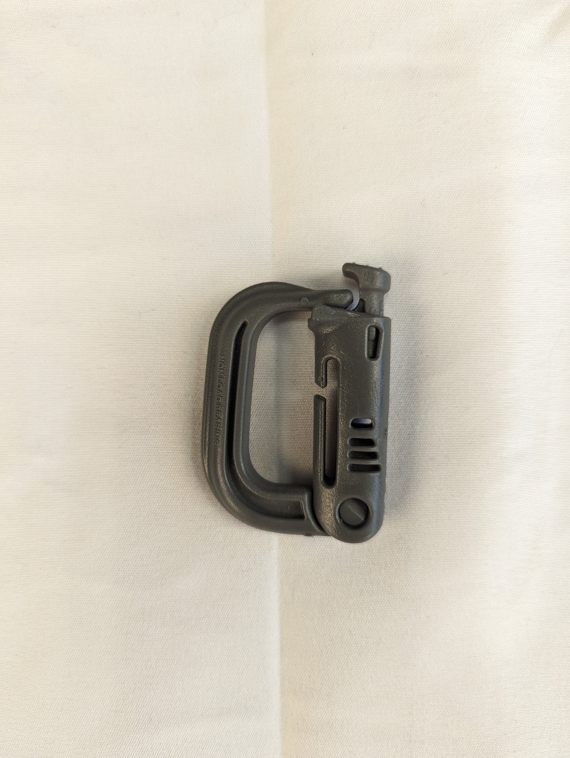 A close-up studio shot of a single green ITW Nexus MOLLE Grimloc D-ring carabiner clip resting diagonally on a wrinkled white fabric background.