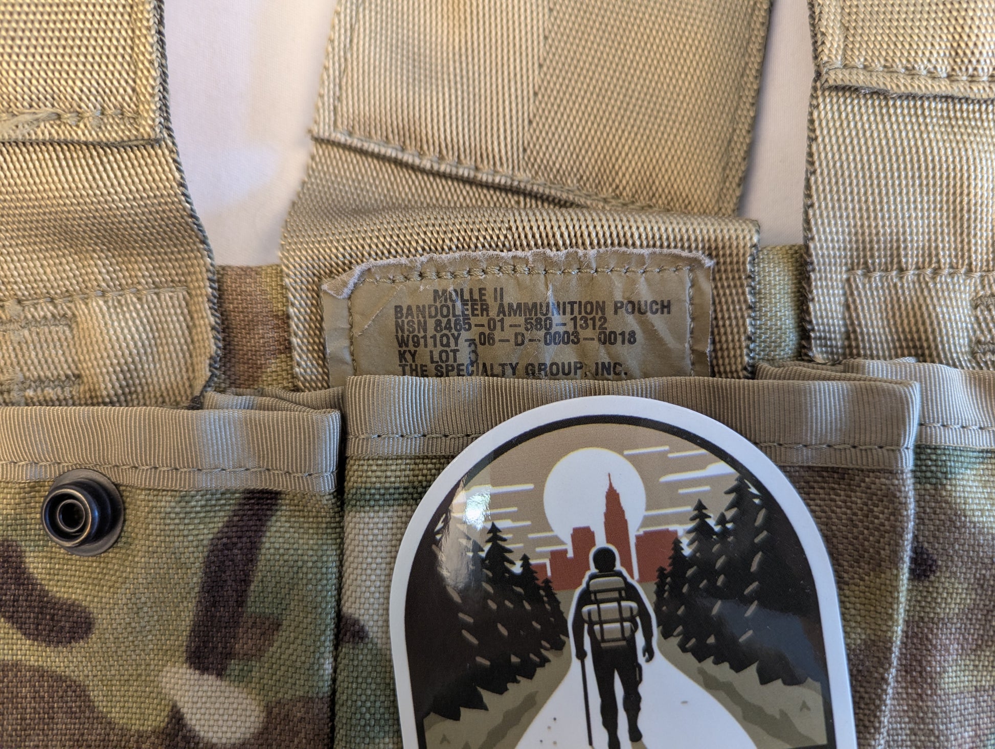 A close-up of a U.S. Military OCP/Multicam Bandoleer pouch, featuring the official NSN issue tag, a snap button closure, and a circular 'FOREST CITY SUPPLY' logo sticker on the flap.