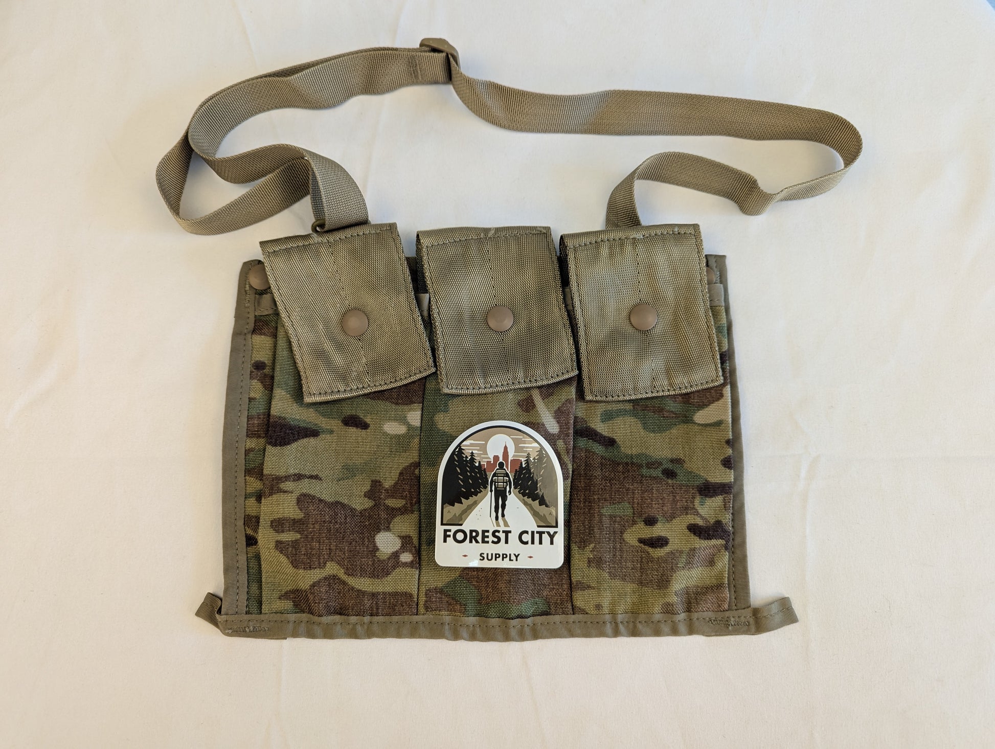 A U.S. Military Surplus OCP/Multicam Bandoleer laid flat, featuring three snap-button magazine pockets, a khaki strap, and a prominent 'FOREST CITY SUPPLY' logo sticker on the front.