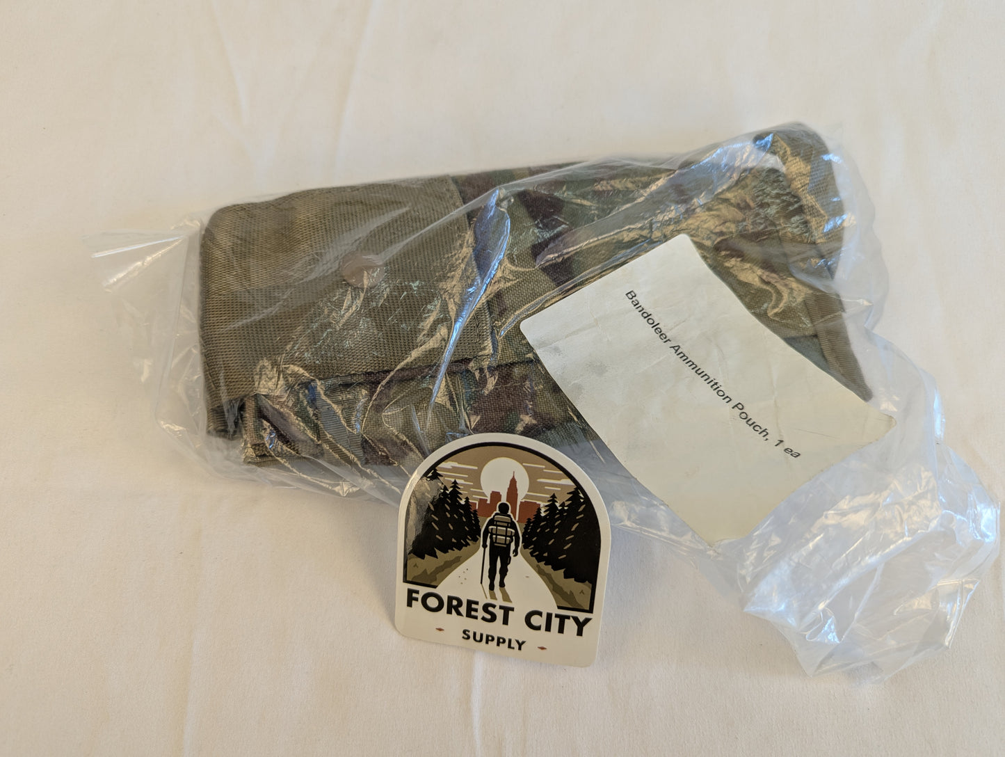 A new U.S. Military Surplus OCP/Multicam Bandoleer pouch sealed inside clear plastic packaging, featuring the original white issue label and a large 'FOREST CITY SUPPLY' logo sticker on the front.