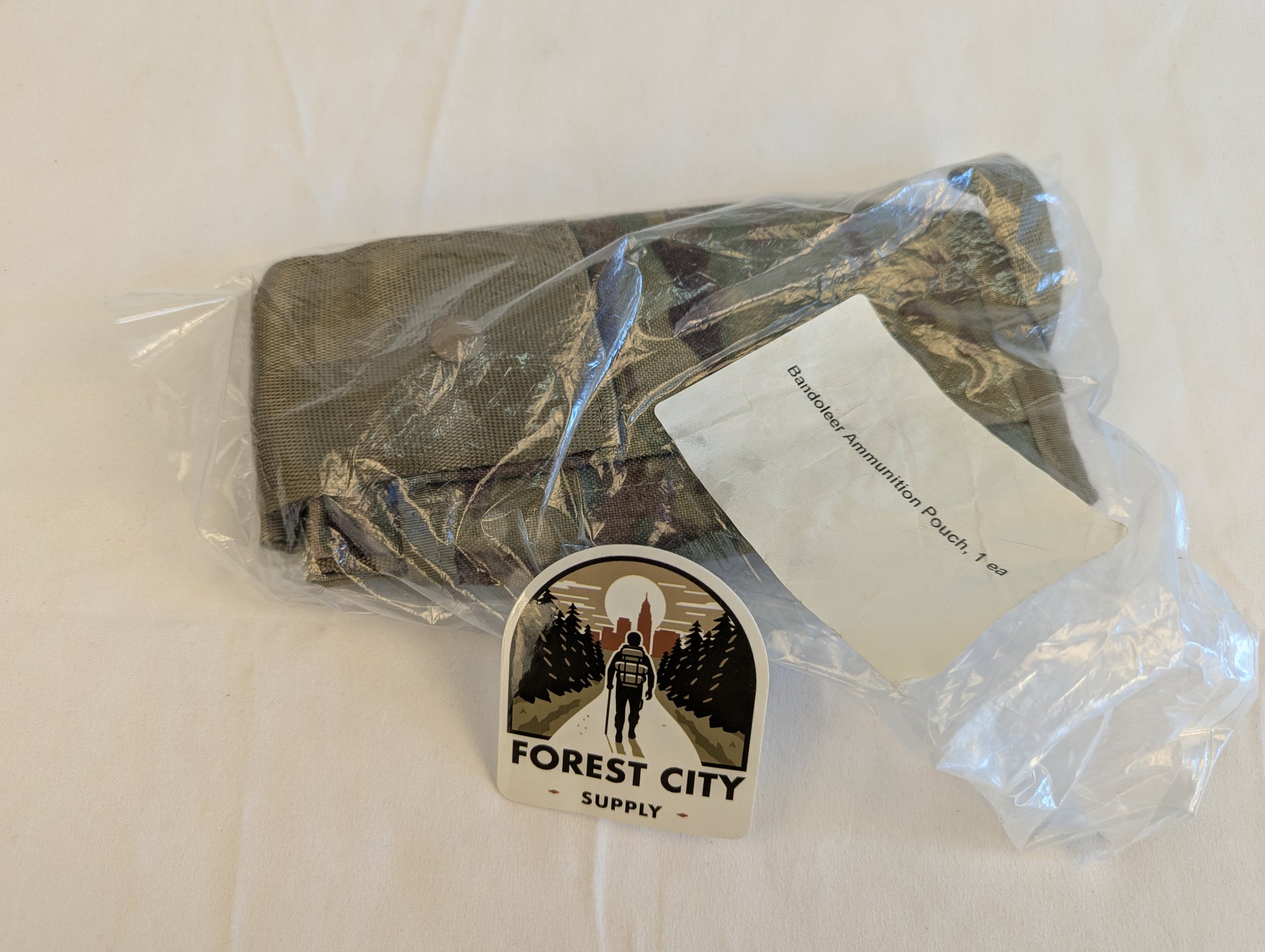 A new U.S. Military Surplus OCP/Multicam Bandoleer pouch sealed inside clear plastic packaging, featuring the original white issue label and a large 'FOREST CITY SUPPLY' logo sticker on the front.