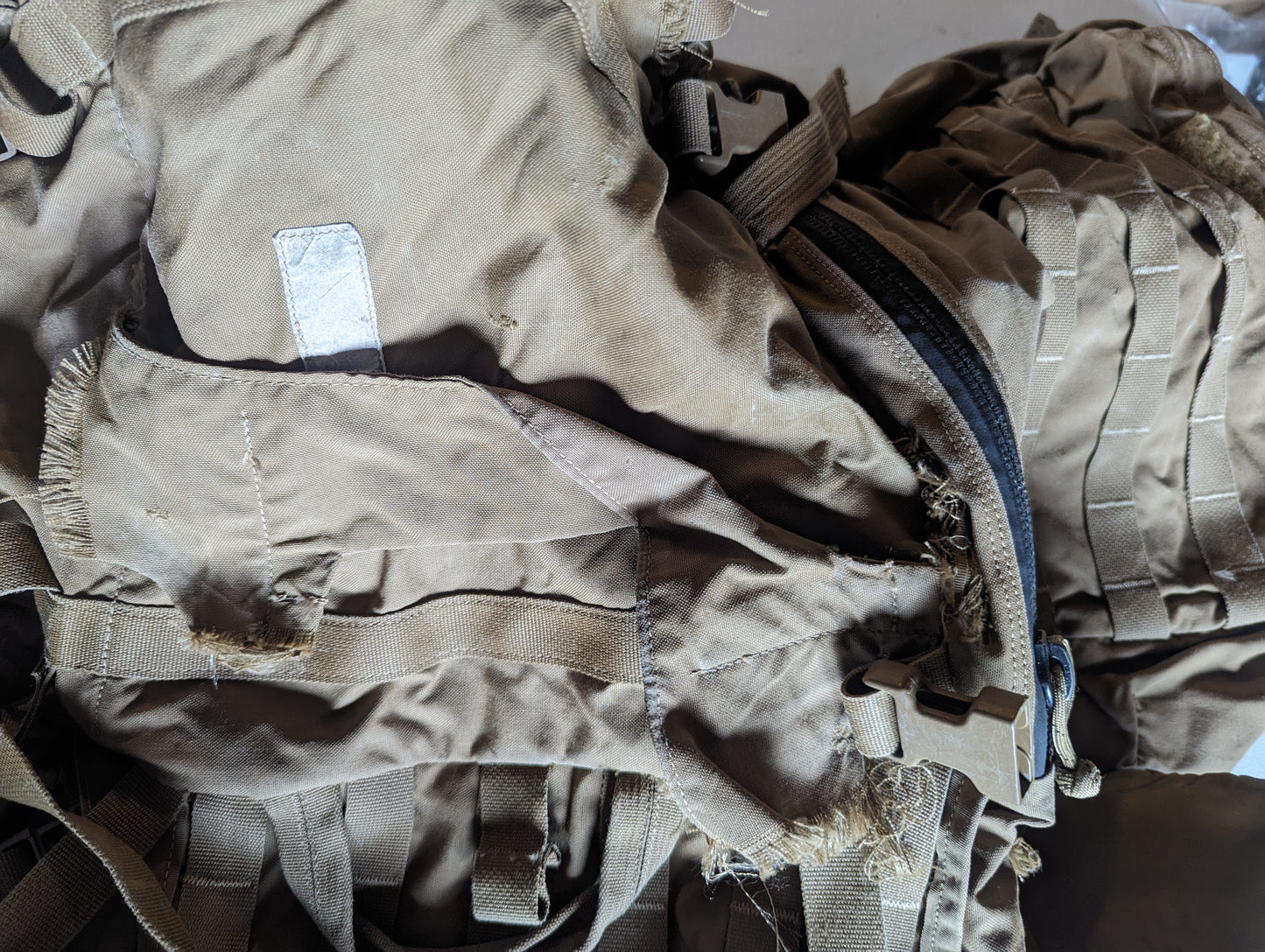 An extreme close-up of a damaged Coyote Brown tactical rucksack, highlighting severe fraying and tearing of the nylon fabric near a zipper and buckle, documenting extensive surplus wear.