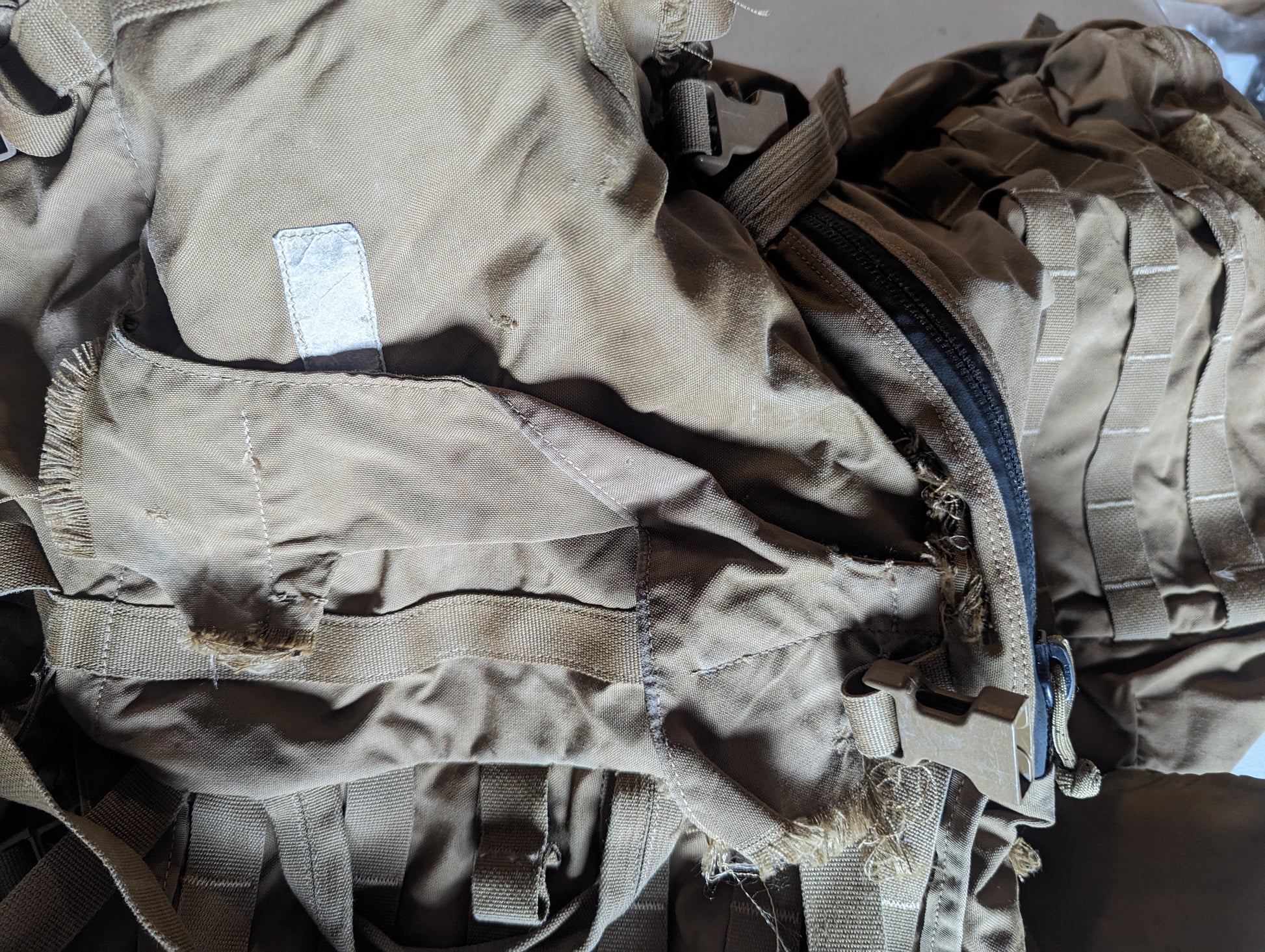 An extreme close-up of a damaged Coyote Brown tactical rucksack, highlighting severe fraying and tearing of the nylon fabric near a zipper and buckle, documenting extensive surplus wear.