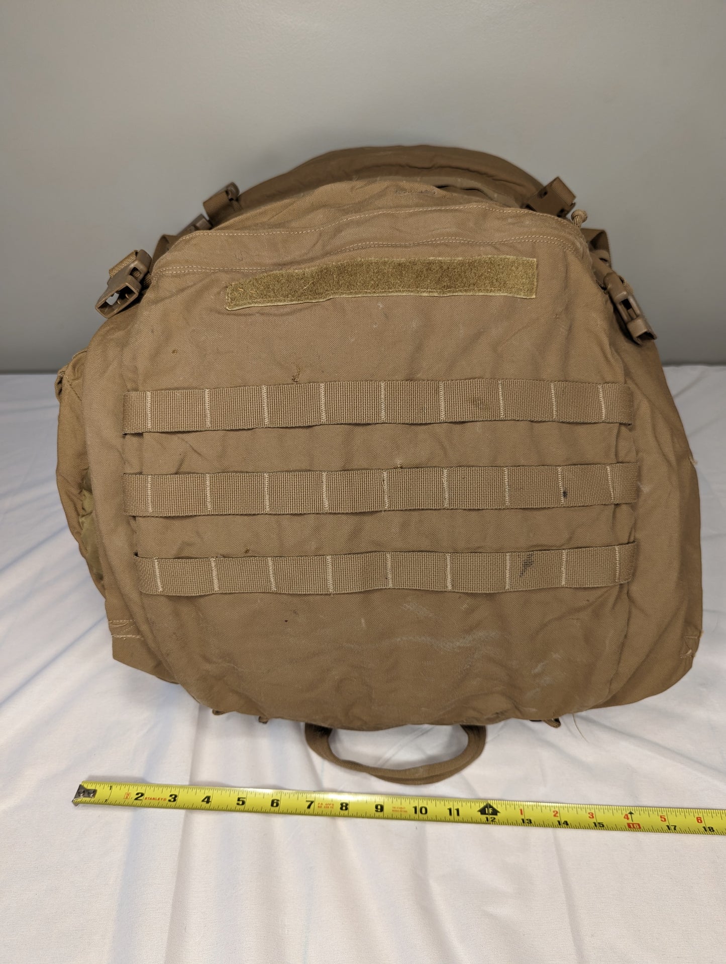 A bottom view of a U.S. Military Surplus USMC FILBE Rucksack in Coyote Brown, lying flat on a white background, with a yellow tape measure placed across the base to demonstrate the width.