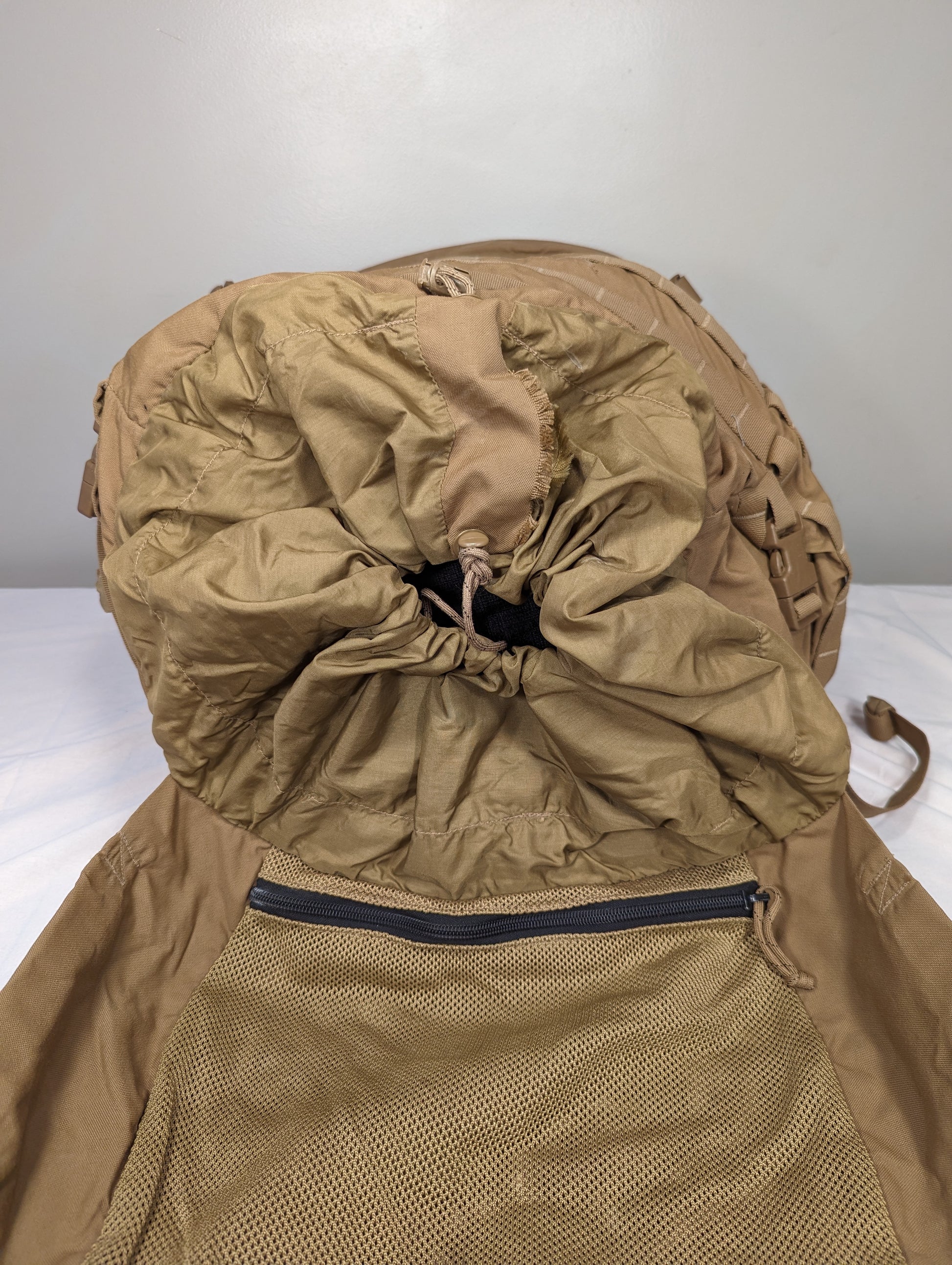 A close-up view of the main compartment opening of a U.S. Military USMC FILBE Rucksack in Coyote Brown, highlighting the tightly cinched drawstring storm collar and a small zippered pocket on the pack body.