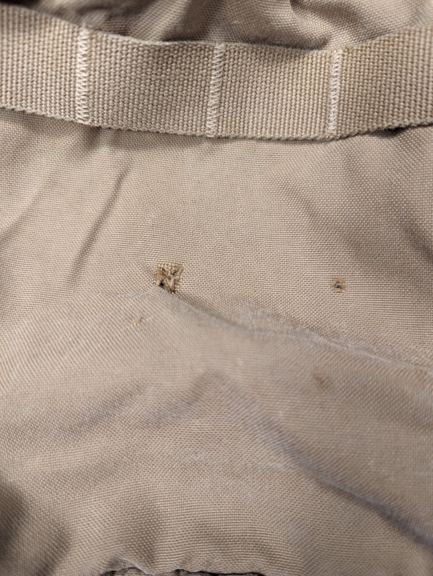 An extreme close-up of light brown military pack fabric, highlighting two small snags or holes below a horizontal webbing strap, documenting the used/defect condition.