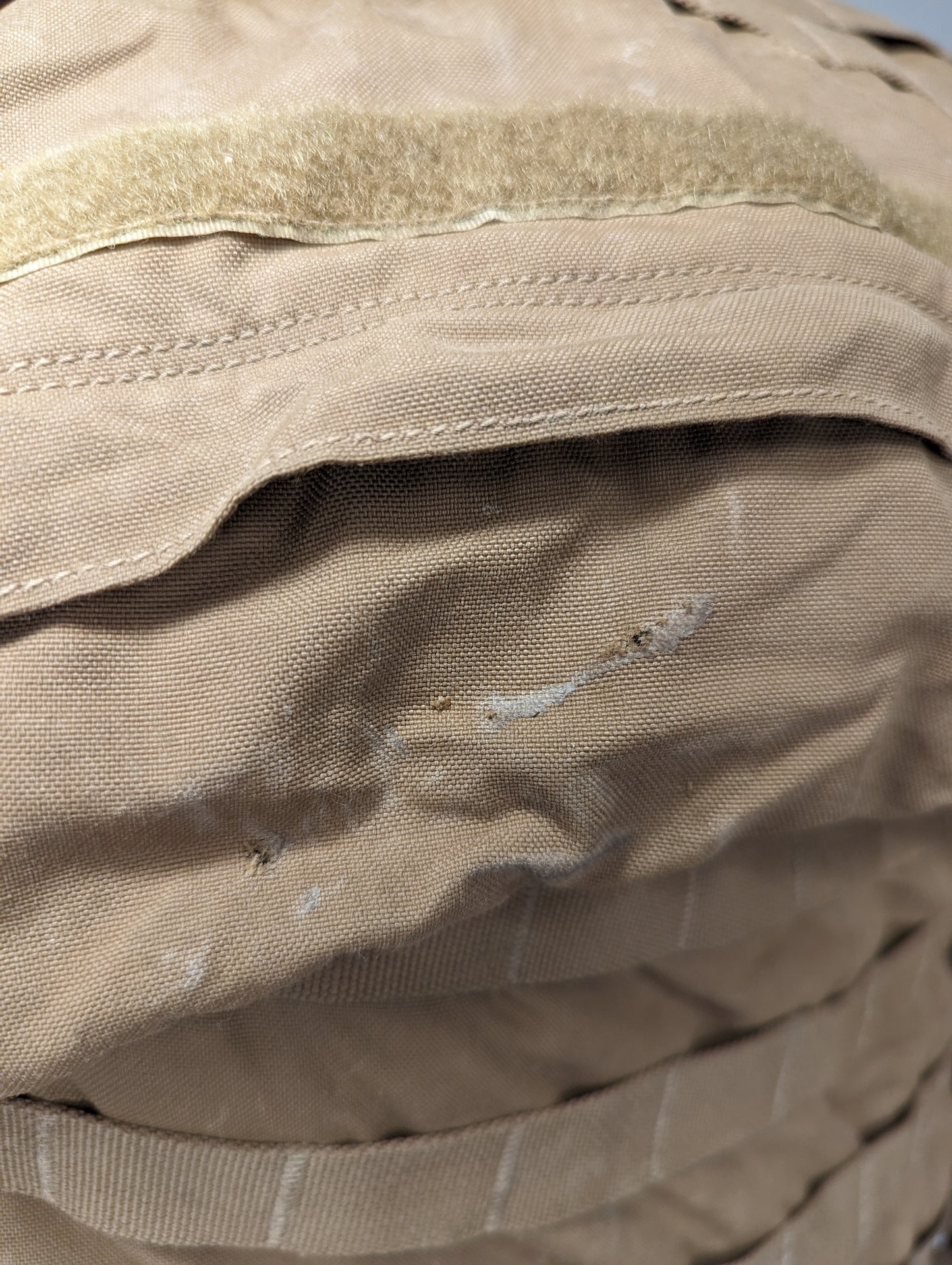A close-up detail shot of the Coyote Brown fabric on a USMC FILBE Rucksack, showing a visible tear, abrasion, and residue marks on the exterior pocket, indicating surplus condition.