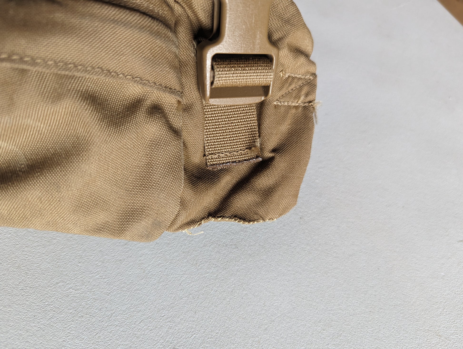 An extreme close-up detail shot of a Coyote Brown tactical pack, showing a tan plastic quick-release buckle attached to webbing, with the fabric heavily frayed and damaged at the bottom edge.