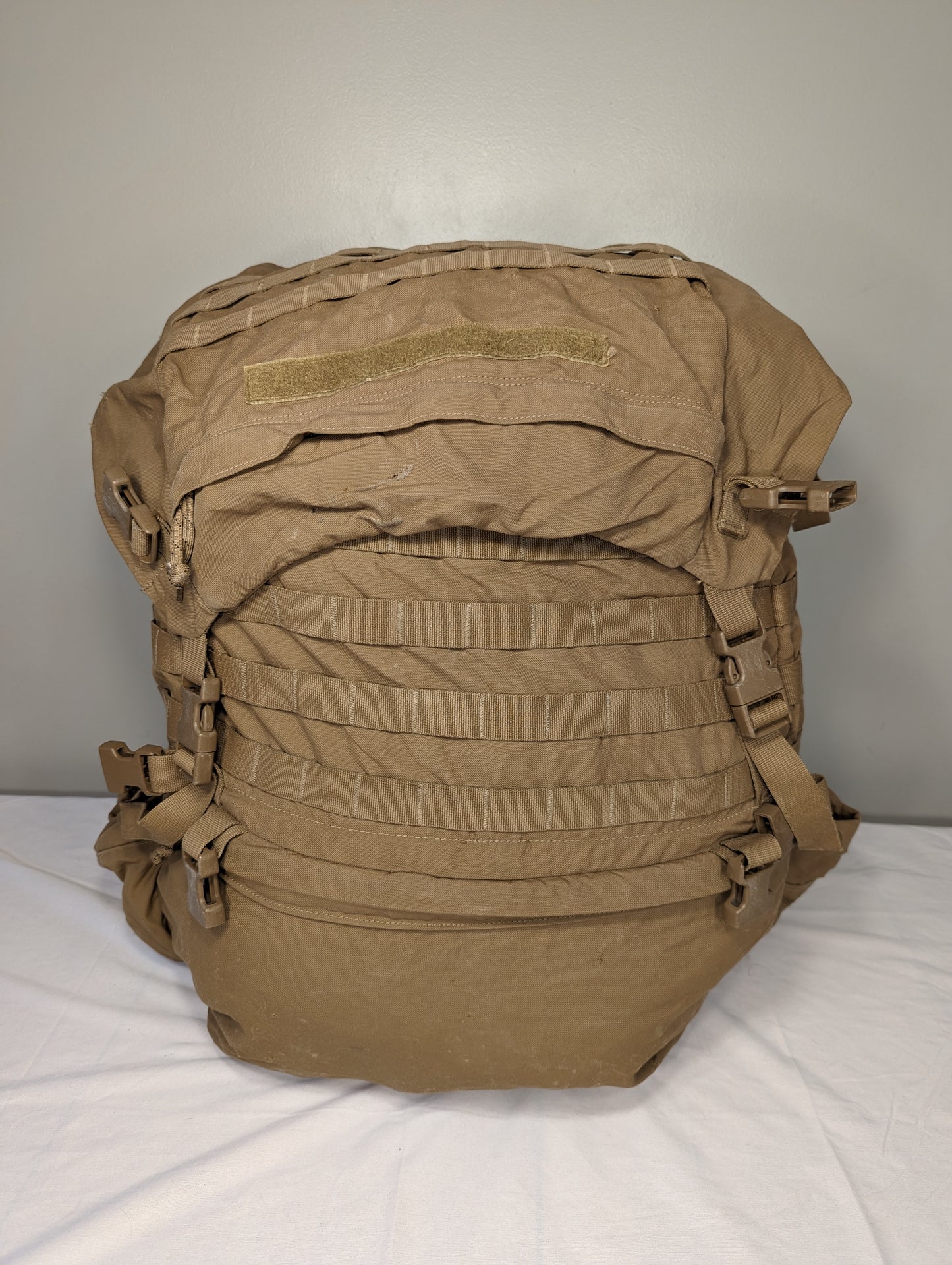 A front view of a U.S. Military Surplus USMC FILBE Rucksack in Coyote Brown, standing upright on a white surface, featuring rows of MOLLE webbing, side compression straps, and a top flap with a hook-and-loop field.