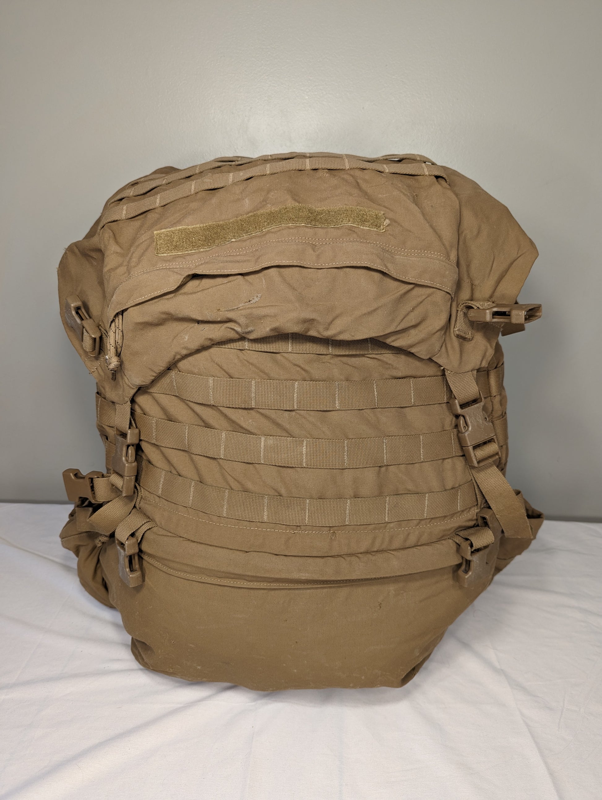 A front view of a U.S. Military Surplus USMC FILBE Rucksack in Coyote Brown, standing upright on a white surface, featuring rows of MOLLE webbing, side compression straps, and a top flap with a hook-and-loop field.
