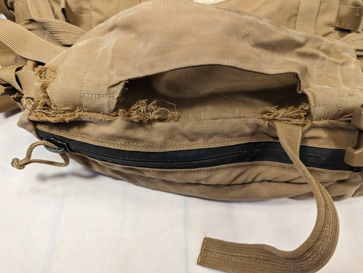 A close-up detail shot of a U.S. Military Coyote Brown pack, showing a large, heavily frayed rip and tear in the nylon fabric near a black zipper, documenting a severe surplus defect.