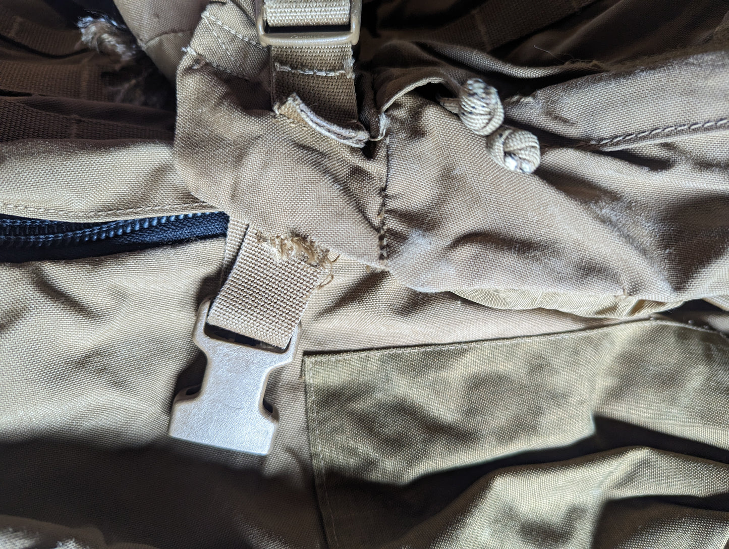 An extreme close-up detail shot of the Coyote Brown fabric on a USMC FILBE Rucksack, highlighting a severe tear and fraying near the main zipper, with a tan quick-release plastic buckle prominently visible.