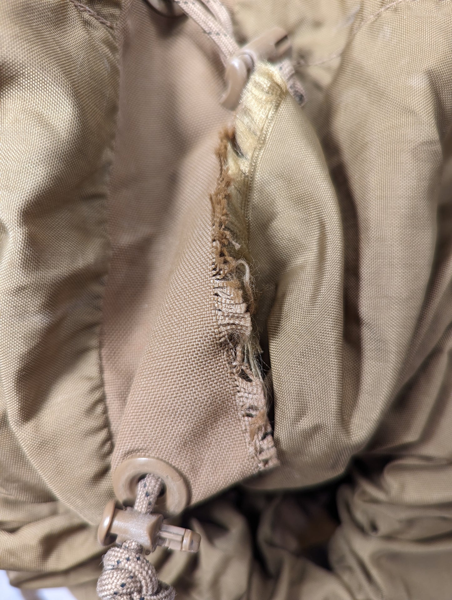 An extreme close-up photograph of a U.S. Military Surplus Coyote Brown pack, highlighting a severe, heavily frayed tear in the fabric near a drawstring channel and cord lock.