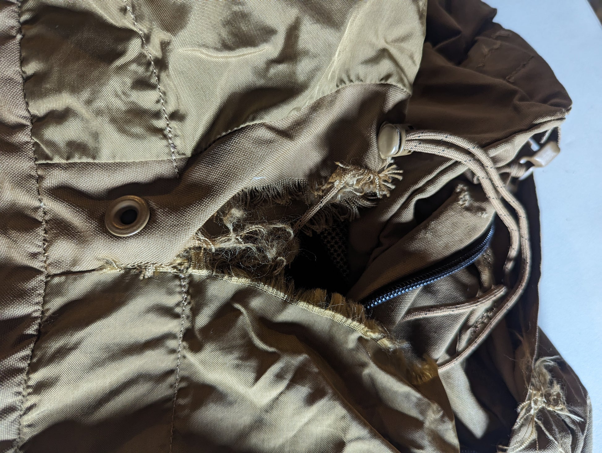 An extreme close-up detail shot of the U.S. Military Coyote Brown pack, showing severe fraying and a large rip around a metal grommet and zipper opening, documenting significant fabric damage and wear.