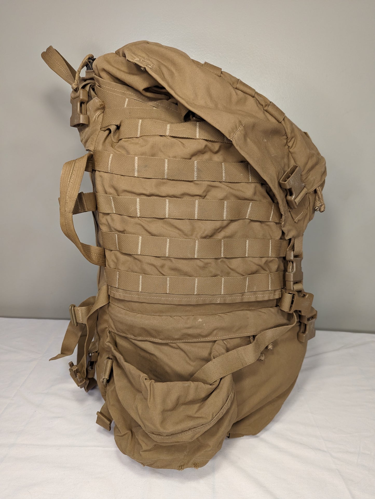 A side view of a U.S. Military Surplus USMC FILBE Rucksack in Coyote Brown, standing upright on a white surface, displaying the MOLLE webbing rows, side compression straps, and sturdy buckle closures.