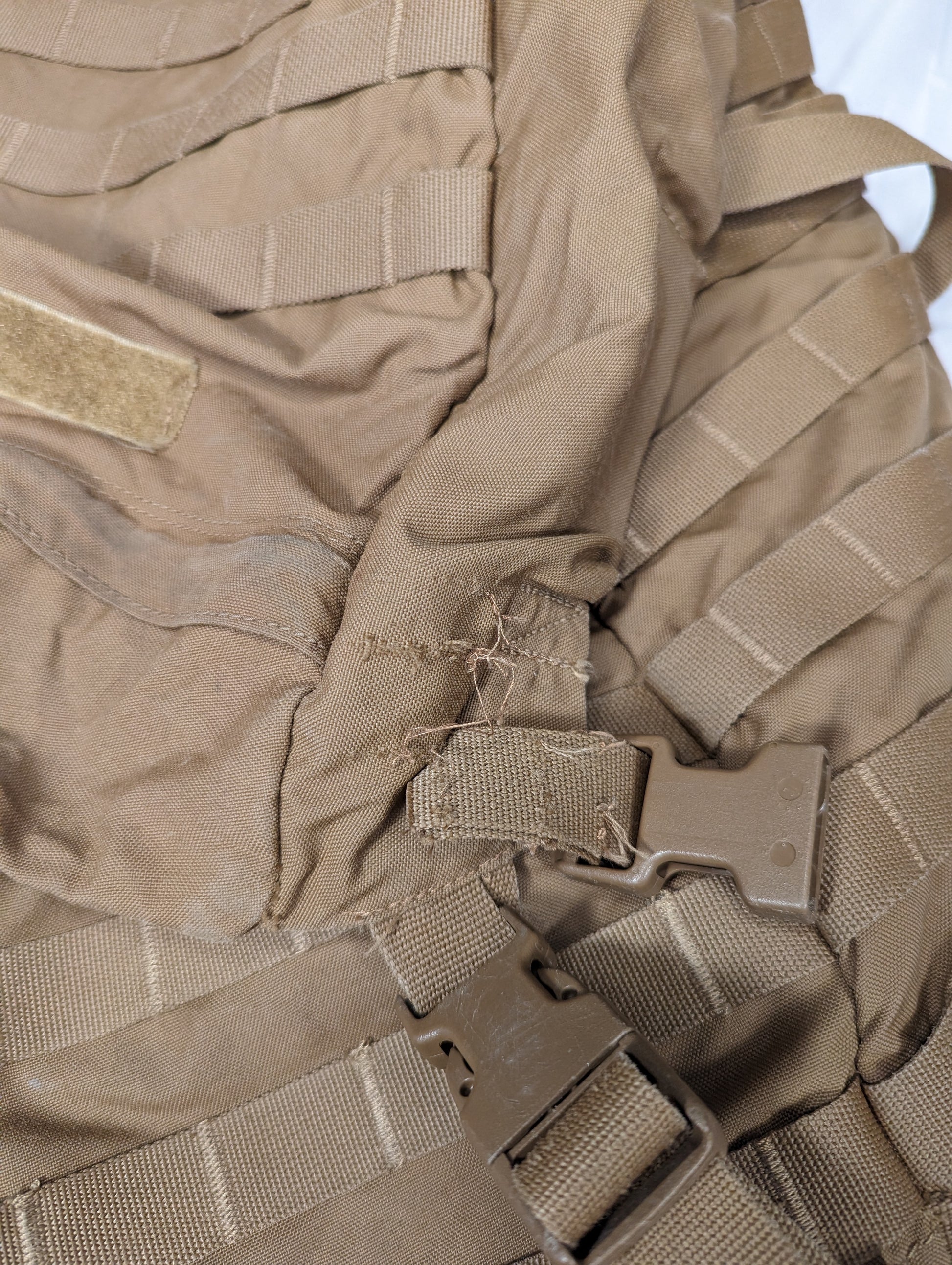 An extreme close-up of the Coyote Brown fabric of a U.S. Military surplus pack, showing a significant tear and broken stitching at a critical webbing and plastic buckle attachment point.