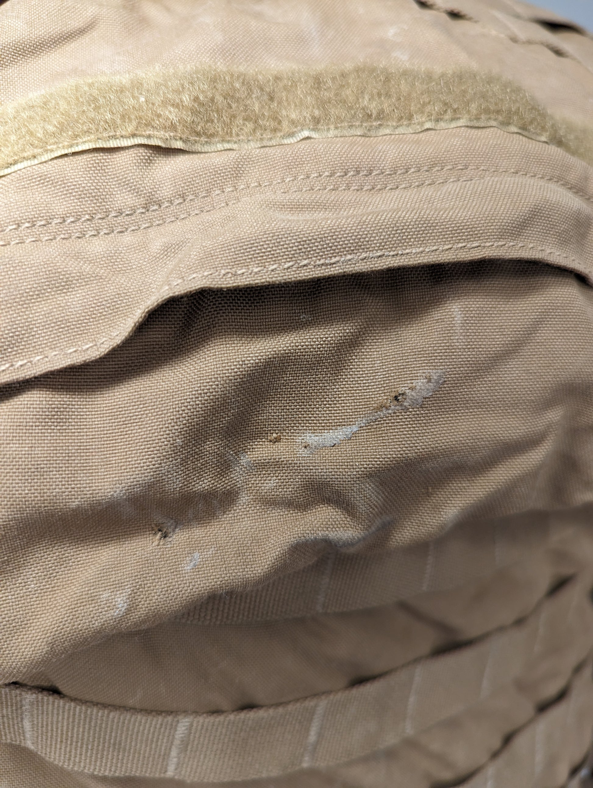 A close-up photograph of the tan nylon fabric on a U.S. Military pack, highlighting a distinct tear and heavy scuffing just below a horizontal Velcro strip, indicating heavy use and damage.