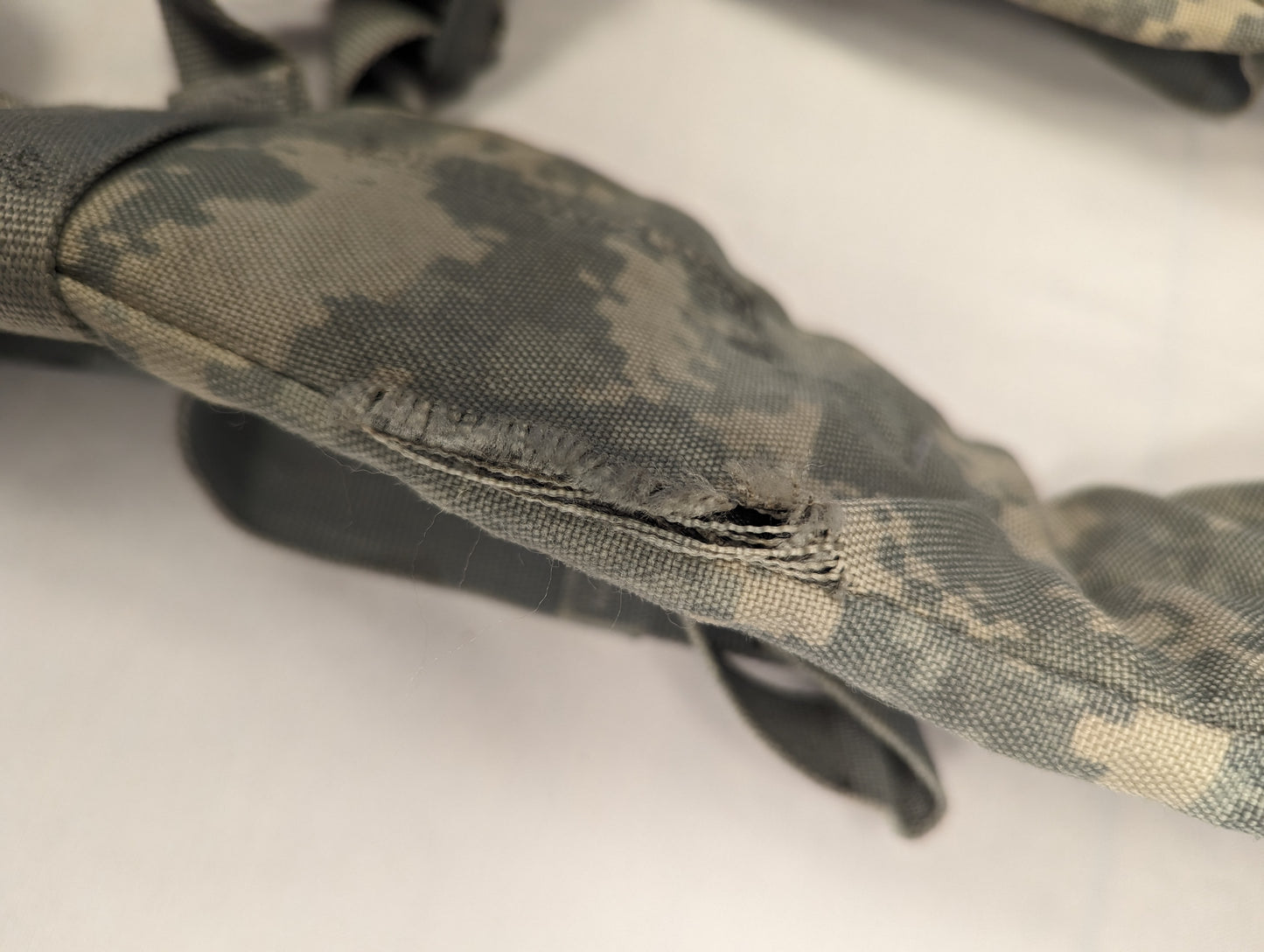 An extreme close-up photograph of a tear and heavy fraying in the ACU digital camouflage fabric of an Enhanced Frame Shoulder Strap, documenting a specific surplus defect.