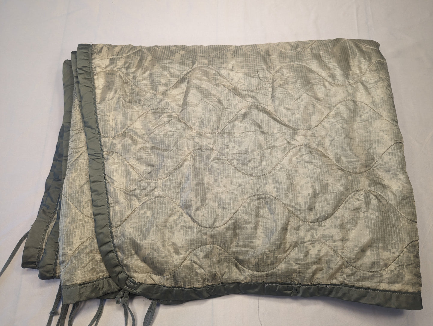 A U.S. Military Surplus Poncho Liner (Woobie) in solid sage green, folded, displaying the distinctive wavy quilted stitch pattern, dark binding, and attachment tie cords.