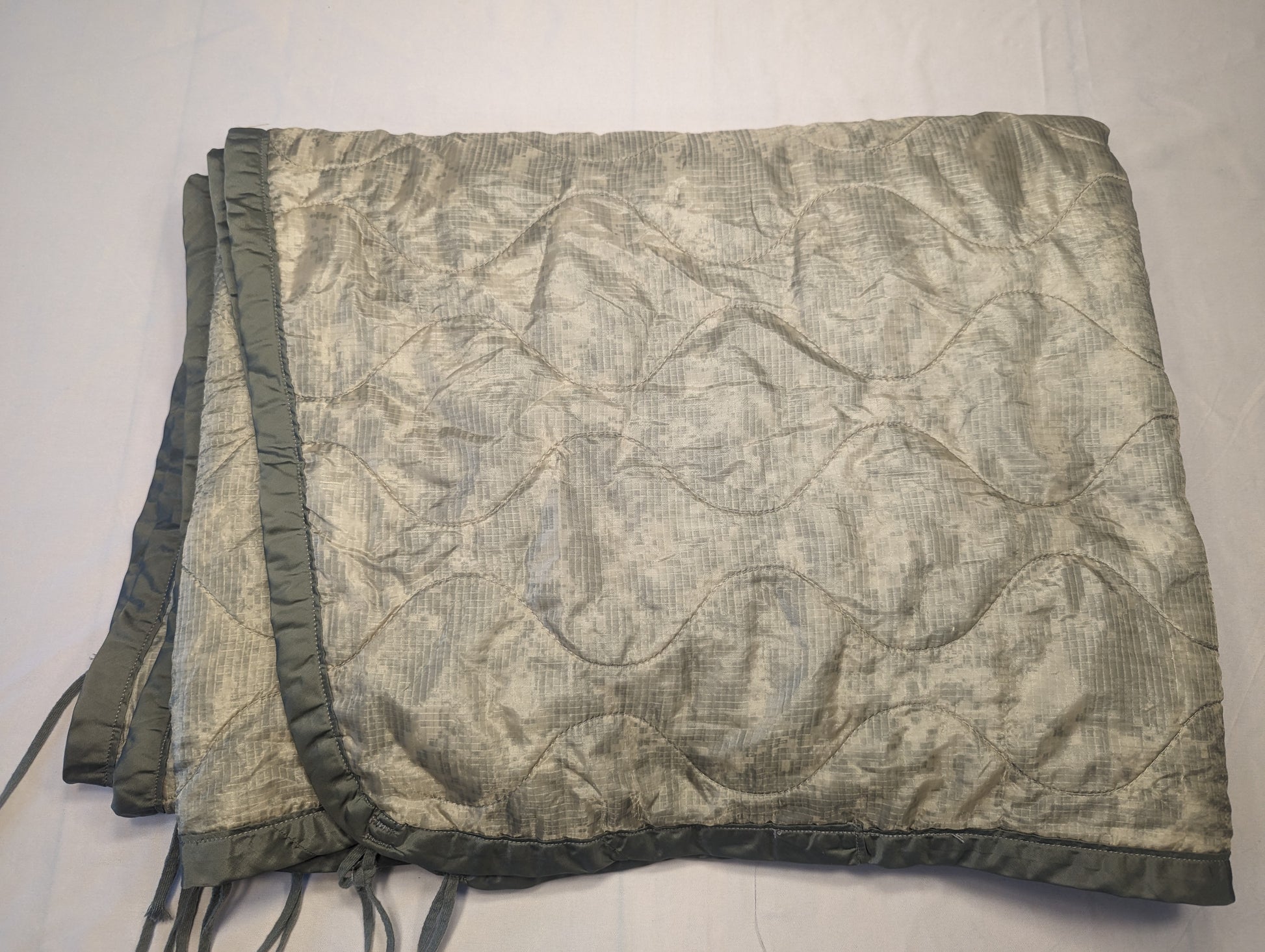 A U.S. Military Surplus Poncho Liner (Woobie) in solid sage green, folded, displaying the distinctive wavy quilted stitch pattern, dark binding, and attachment tie cords.